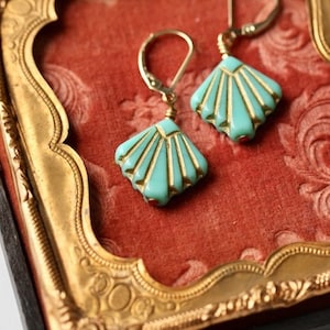 May include: A pair of turquoise blue and gold earrings with a fan-shaped design. The earrings are displayed in a velvet-lined box with a gold frame.