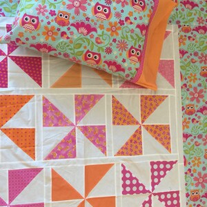 May include: A white quilt with a patchwork design featuring pink, orange, and white pinwheels. The quilt is decorated with a pillow featuring a blue background with a repeating pattern of owls, flowers, and birds.