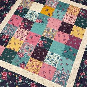 May include: A patchwork quilt with a variety of floral and paisley patterns in shades of pink, teal, purple, and yellow. The quilt is surrounded by a white border and a dark blue border with floral designs.