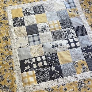 May include: A patchwork quilt with a variety of fabric patterns. The quilt features squares of floral, plaid, and dotted fabrics in shades of yellow, navy, gray, and white. The border is a yellow floral print.
