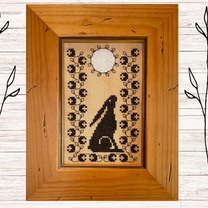 Könnte beinhalten: Eine gerahmte Kreuzstich-Stickerei mit einer schwarzen Silhouette eines Kaninchens unter einem Vollmond. Das Design ist von einem Rand aus schwarzen Blättern und Blumen umgeben.