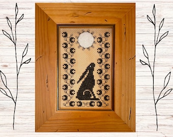 May include: A framed cross-stitch embroidery featuring a black silhouette of a rabbit under a full moon. The design is surrounded by a border of black leaves and flowers.