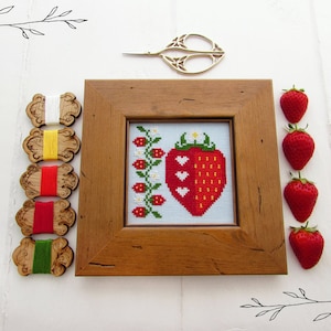 May include: A framed cross-stitch embroidery of a red strawberry with white hearts and green leaves. The embroidery is surrounded by a wooden frame and is displayed on a white surface with wooden spools of embroidery floss and red strawberries.