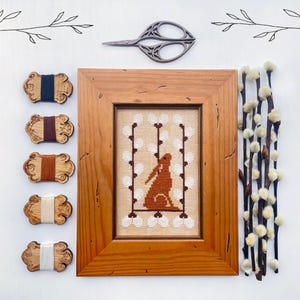 May include: A framed cross-stitch of a brown rabbit, surrounded by white polka dots, with a wooden frame. Wooden thread bobbins with various coloured threads are arranged to the left. A pair of scissors and pussy willow branches are also present.