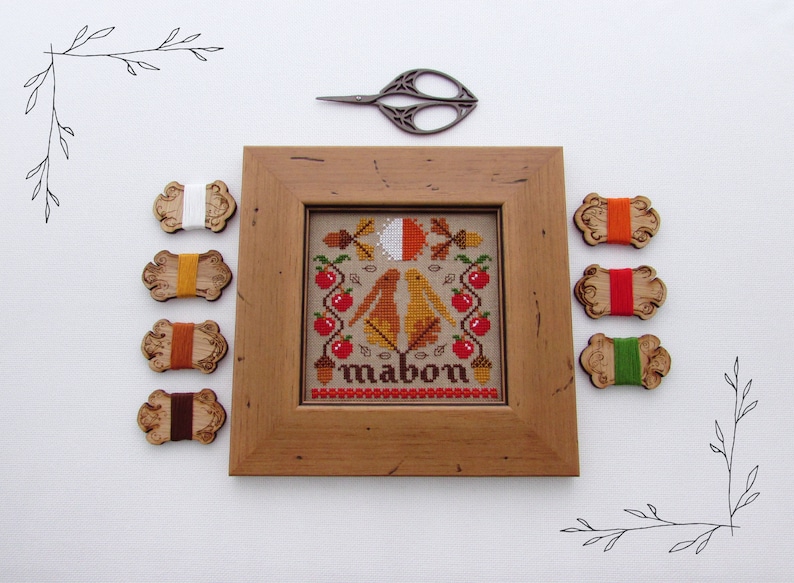 May include: A framed cross-stitch embroidery with a fall theme. The design features two birds, leaves, acorns, and apples. The word "Mabon" is stitched below the design. The embroidery is surrounded by wooden spools of embroidery floss in various colors.