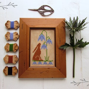 May include: A framed cross-stitch embroidery featuring a brown rabbit sitting near a cluster of blue bell flowers. The embroidery is displayed in a wooden frame with a brown finish. The image also includes wooden spools of thread in various colors.