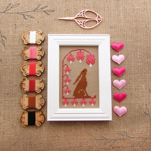 May include: A white frame containing a cross-stitch design of a brown rabbit sitting under a canopy of pink flowers. The frame is surrounded by wooden spools of embroidery floss and pink felt hearts.