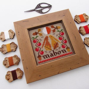 May include: A framed cross-stitch embroidery with a fall theme. The design features two birds, apples, leaves, and acorns. The word "Mabon" is stitched in the center. The embroidery is surrounded by a wooden frame and there are several spools of embroidery floss in various colors.