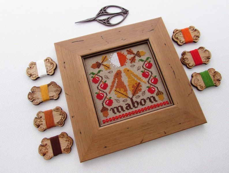 May include: A framed cross-stitch embroidery with a fall theme. The design features two birds, apples, leaves, and acorns. The word "Mabon" is stitched in the center. The embroidery is surrounded by a wooden frame and there are several spools of embroidery floss in various colors.