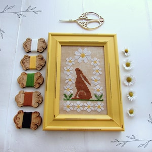 May include: A yellow frame with a cross-stitch design of a brown rabbit sitting in a wreath of white daisies. The frame is on a white surface with wooden spools of embroidery floss and a pair of gold scissors.