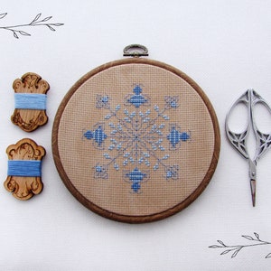 May include: A blue and white cross-stitch design of a snowflake in a wooden embroidery hoop with two wooden thread bobbins and silver embroidery scissors on a white background.