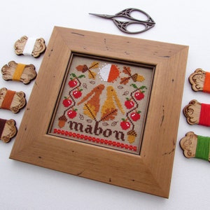 May include: A framed cross-stitch embroidery with a fall theme. The design features two birds, apples, leaves, and acorns. The word "Mabon" is stitched in brown thread at the bottom of the design. The embroidery is surrounded by a wooden frame and there are eight spools of embroidery floss in various colors.