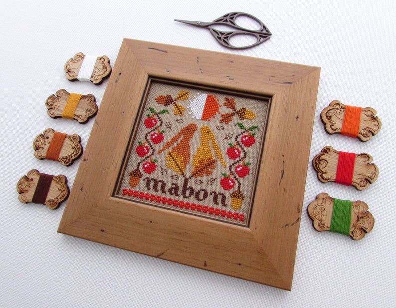 May include: A framed cross-stitch embroidery with a fall theme. The design features two birds, apples, leaves, and acorns. The word "Mabon" is stitched in brown thread at the bottom of the design. The embroidery is surrounded by a wooden frame and there are eight spools of embroidery floss in various colors.