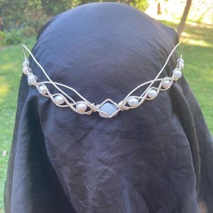 Könnte beinhalten: Ein silberfarbener Draht-Haarschmuck mit einem hellblauen Edelstein in der Mitte und abwechselnd weißen Perlen. Der Haarschmuck hat ein zartes, handgefertigtes Aussehen, geeignet für besondere Anlässe oder Kostüme.