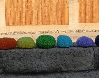 May include: A row of seven colorful, crocheted balls in shades of pink, orange, yellow-green, green, blue, purple, and navy blue. The balls are arranged on a gray stone surface, with a wooden fence and a building in the background.