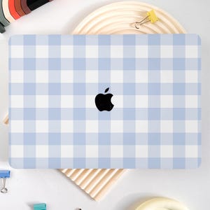 Peut inclure: Un ordinateur portable avec un motif vichy bleu clair et blanc. Il arbore un logo Apple noir au centre. Il est entouré de fournitures de bureau colorées, dont des pinces et un arc-en-ciel en bois.