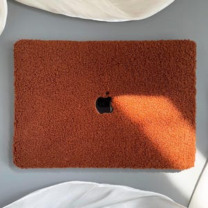 Könnte beinhalten: Eine braune, flauschige Laptop-Hülle mit einem Apple-Logo-Ausschnitt.