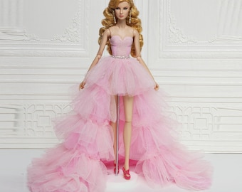 May include: A pink tulle dress with a high-low hemline. The dress has a fitted bodice with a sweetheart neckline and a sparkly belt. The doll is wearing red heels.
