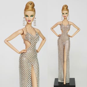 May include: Two fashion dolls wearing silver chainmail dresses. The dolls have blonde hair and are wearing silver earrings. The doll on the left is standing with her arms at her sides. The doll on the right is standing with her arms crossed.