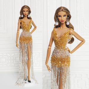 May include: Two fashion dolls wearing long, beaded gowns with gold and silver accents. The gowns have a sheer bodice and a fringe skirt. The dolls have long, dark hair and are wearing pearl earrings.