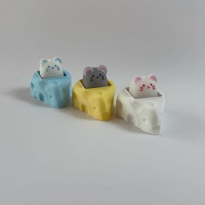 May include: Three ceramic mouse figurines in cheese-shaped holders. The mice are light blue, gray, and pink, each with small dot details. The cheese holders are light blue, yellow, and white, with holes. The figurines are on a white surface.
