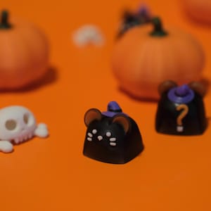 Peut inclure: Des touches de clavier en forme de souris noires sur le thème d'Halloween, avec des chapeaux de sorcière violets et des accents blancs. Les touches sont posées sur un fond orange avec des citrouilles floues et un crâne.