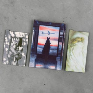 May include: Three rectangular flat items featuring illustrations. The left shows a figure holding a small child in a window. The center depicts a silhouette looking at an airplane against a sunset. The right shows a figure sleeping under a white blanket.
