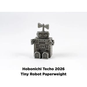 Könnte beinhalten: Ein kleiner, metallischer Roboter-Briefbeschwerer mit dem Text "YOU LOVE YOU" auf seinem Körper. Der Roboter hat einen quadratischen Körper, runde Augen und eine propellerartige Antenne. Auf der Basis steht "Hobonichi Techo 2026 Tiny Robot Paperweight."