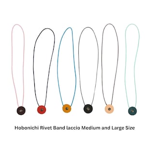Op de afbeelding: Zes Hobonichi Rivet Band laccio accessoires in medium en large formaat. Elk heeft een dun elastisch koord in roze, zwart, blauw, zwart, beige of mintgroen, bevestigd aan een ronde lederachtige tab met een centrale metalen klinknagel.