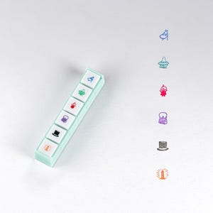 Pode incluir: Um conjunto de seis carimbos quadrados individuais numa caixa retangular verde-menta claro. Cada carimbo apresenta um ícone único numa cor diferente: uma personagem azul, um chapéu azul-petróleo, uma figura vermelha, uma mala roxa, uma cartola preta e um farol laranja.