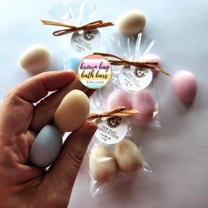 May include: A hand holding two egg-shaped soaps, one light beige and one light blue. Several other egg-shaped soaps in various pastel colours are packaged in clear cellophane bags, tied with raffia and small tags. The text "brown bag bath bars" is visible.