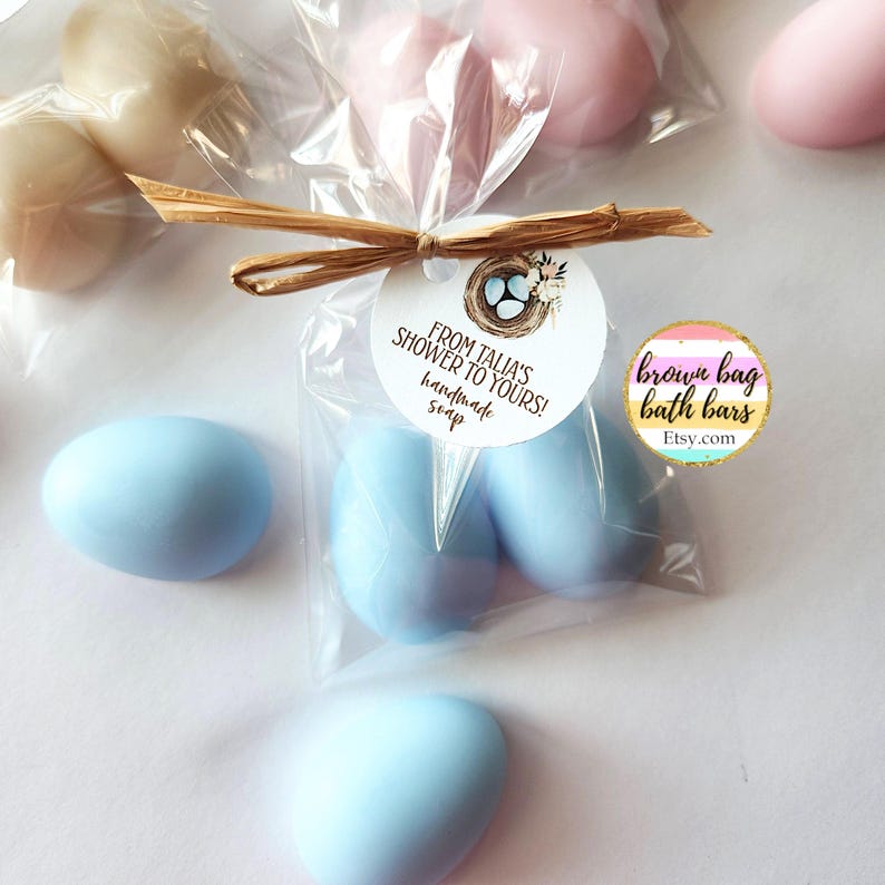 May include: Egg-shaped handmade soaps in blue, pink, and beige, packaged in clear cellophane bags. Each bag is tied with raffia and a circular tag that reads "From Talia's Shower to Yours!" and "handmade soap". A brown bag bath bars Etsy.com sticker is visible.