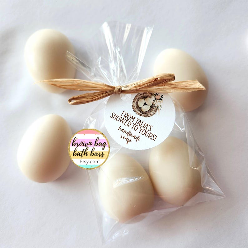 May include: Egg-shaped handmade soaps in a clear cellophane bag tied with raffia. A round tag reads "From Talia's Shower to Yours! handmade soap." A second tag says "brown bag bath bars" with an Etsy.com URL.