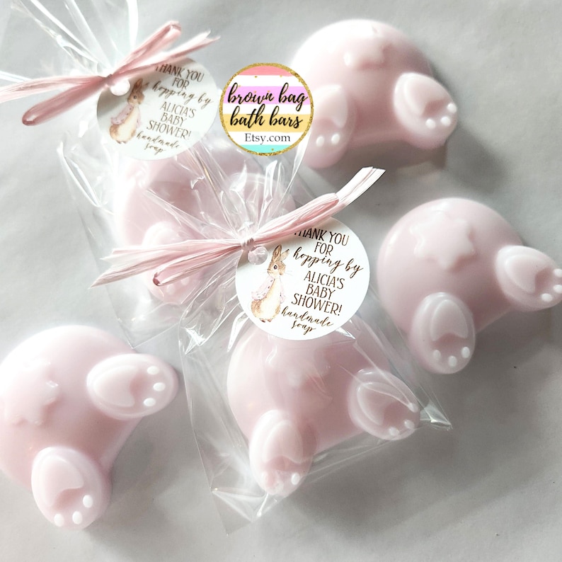 K&ouml;nnte beinhalten: Rosa, hasenf&ouml;rmige Seifen, verpackt in transparenten Zellophanbeuteln und mit rosa Bastb&auml;ndern gebunden. Jede Seife hat wei&szlig;e Pfoten-Details. Die Seifen sind mit "ALICIA'S BABY SHOWER! handmade soap" beschriftet. Der Etsy-Shop-Name "brown bag bath bars" ist ebenfalls sichtbar.