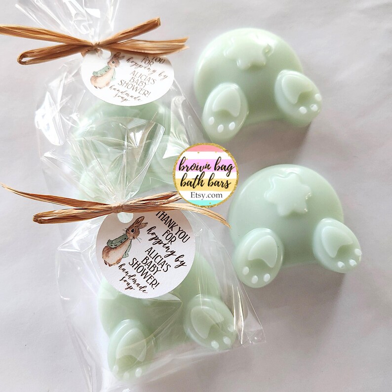 K&ouml;nnte beinhalten: Hellgr&uuml;ne Seifen in Form von Hasenpopo. Zwei Seifen sind einzeln in durchsichtigem Zellophan mit Raffiab&auml;ndern und Etiketten verpackt, auf denen steht: "Vielen Dank f&uuml;r das Hopping bei Alicias Babyparty! handgemachte Seife."