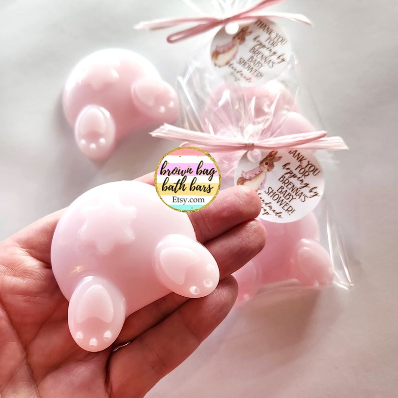 K&ouml;nnte beinhalten: Rosa Seifen in Form eines Hasen-Hinterns, ideal f&uuml;r eine Babyparty. Die Seifen sind einzeln in durchsichtigem Zellophan verpackt und mit rosa Band gebunden. Jede Seife hat ein Etikett mit der Aufschrift "Danke f&uuml;rs Hopping bei Brennas Babyparty!"