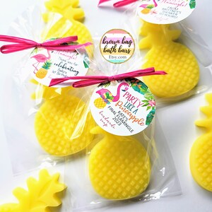May include: Yellow pineapple-shaped soaps in clear cellophane bags, tied with pink raffia bows. Each soap has a textured surface and is adorned with a round tag featuring a flamingo, pineapple, and text. The tags read "Party Like a Pineapple" and "Final Flamingle 2023".