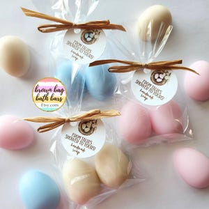 May include: Handmade egg-shaped soaps in pastel shades, packaged in clear cellophane bags and tied with raffia bows. Each bag has a tag that reads "From Talia's Shower to Yours! handmade soap". The soaps are light blue, pink, and cream.