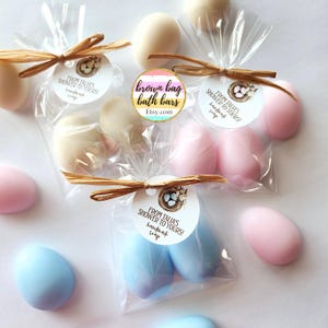 May include: Egg-shaped handmade soaps in cellophane bags, tied with raffia. The soaps are in pastel shades of white, pink, and blue. Each bag has a tag that reads "From Talia's Shower to Yours!" and "handmade soap".