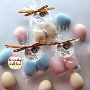 May include: A collection of egg-shaped soaps in pastel shades, such as pink, blue, and cream, are presented in clear cellophane bags, secured with raffia bows. Each bag features a tag that reads "FROM TALIA'S SHOWER TO YOURS! handmade soap."