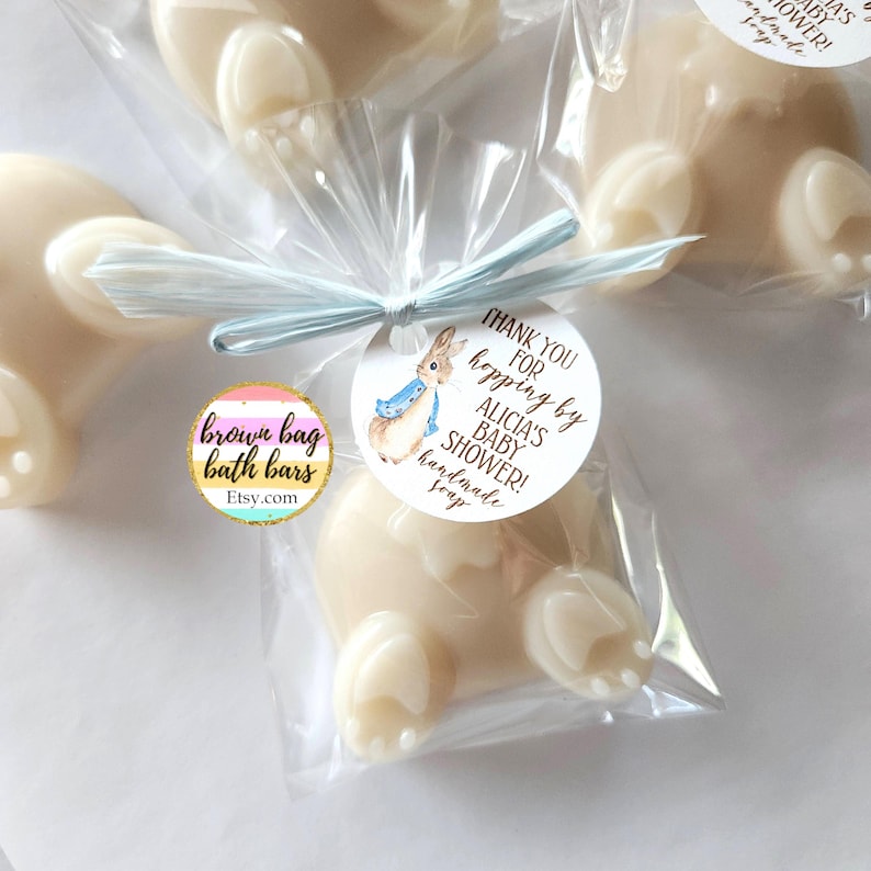 K&ouml;nnte beinhalten: Handgemachte Seifen in Hasenform in transparenten Zellophanbeuteln, gebunden mit hellblauem Band. Jede Seife hat ein Etikett mit einer Peter-Rabbit-Illustration und dem Text "Thank you for hopping by Alicia's Baby Shower!" Die Seife ist von Brown Bag Bath Bars.