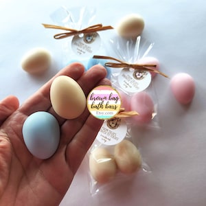 May include: Egg-shaped soaps in pastel shades of cream, blue, and pink. Some are packaged in clear cellophane bags, tied with raffia bows and small tags reading "FROM TALIA'S SHOWER TO YOURS! handmade soap". The Etsy shop name "brown bag bath bars" is also visible.