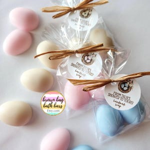 May include: Egg-shaped soaps in pink, cream, and blue are shown. Some are loose, others are packaged in clear bags tied with raffia and tags that read "From Talia's Shower to Yours! Handmade Soap." The logo "brown bag bath bars" is also visible.