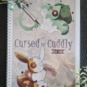 May include: A spiral-bound notebook with the title "Cursed but Cuddly Vol #01". The cover features illustrations of stitched plushie-like creatures, including a green and white Bulbasaur and a brown and white Eevee. The background is light gray with sewing-themed patterns.