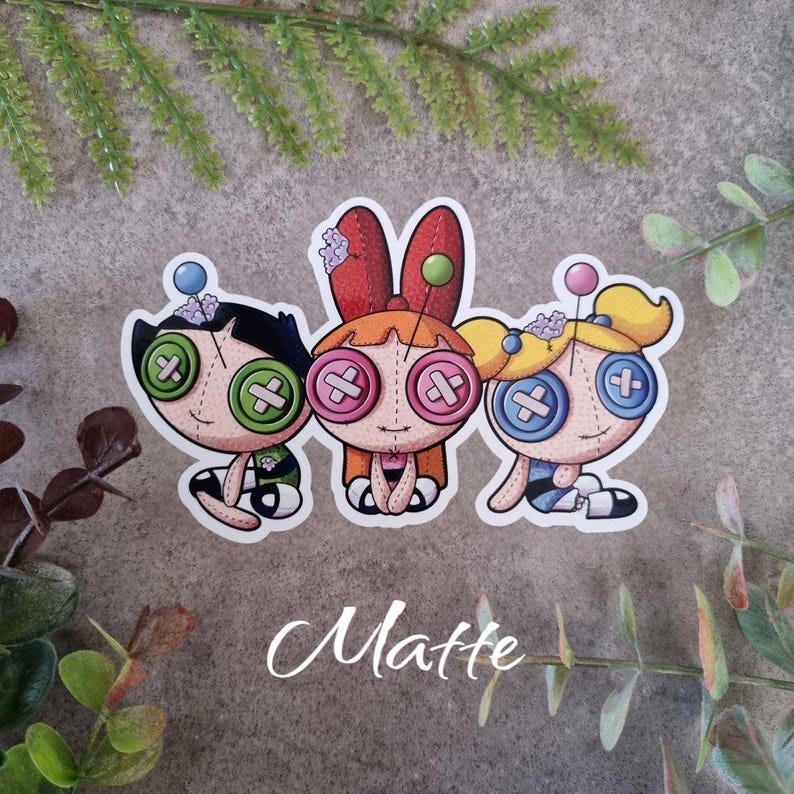 May include: Three cartoon character stickers with button eyes and stitched features. The characters have black, orange, and yellow hair, and are wearing outfits in green, orange, and blue. The word "Matte" is written in cursive at the bottom.