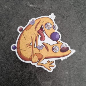 Puede incluir: Una pegatina con un perro y un gato de dibujos animados en una pose juguetona. El perro es de color marrón claro con una nariz y lengua moradas, y el gato es de color marrón claro. La pegatina tiene un borde blanco y está sobre una superficie gris.