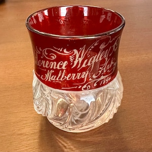 May include: A red glass cup with a clear, textured bottom. The cup has white lettering that reads "Florence Wigley & Mulberry Ark 1894".