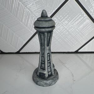May include: A gray, stone-textured sculpture of the Space Needle tower. The object features a tall, slender central column with vertical support beams, a circular observation deck near the top, and a pointed spire, all finished in a weathered gray.