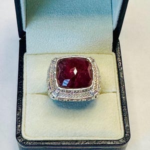 May include: A square-shaped ring featuring a large, deep red gemstone surrounded by a band of smaller, clear gemstones. The ring is set in a silver band with a decorative design.