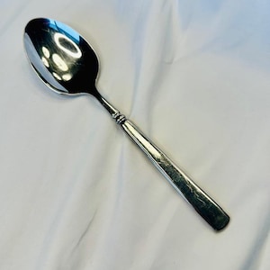 May include: A silver spoon with a rounded bowl and a long, rectangular handle. The spoon reflects the light, showing a shiny surface. The handle has a decorative design near the bowl.